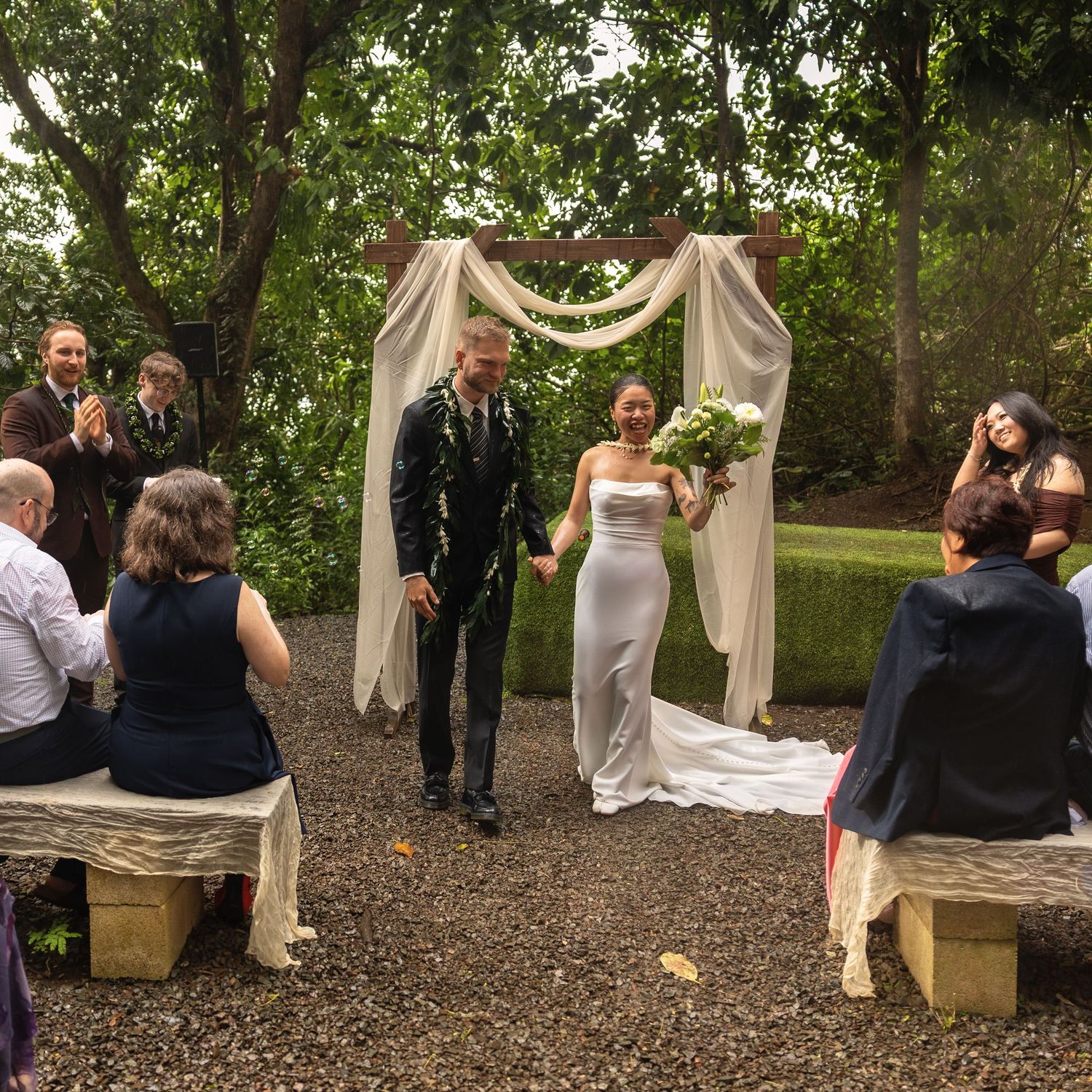 Newlywed couple walks through garden wedding arch while guests applaud their outdoor ceremony.