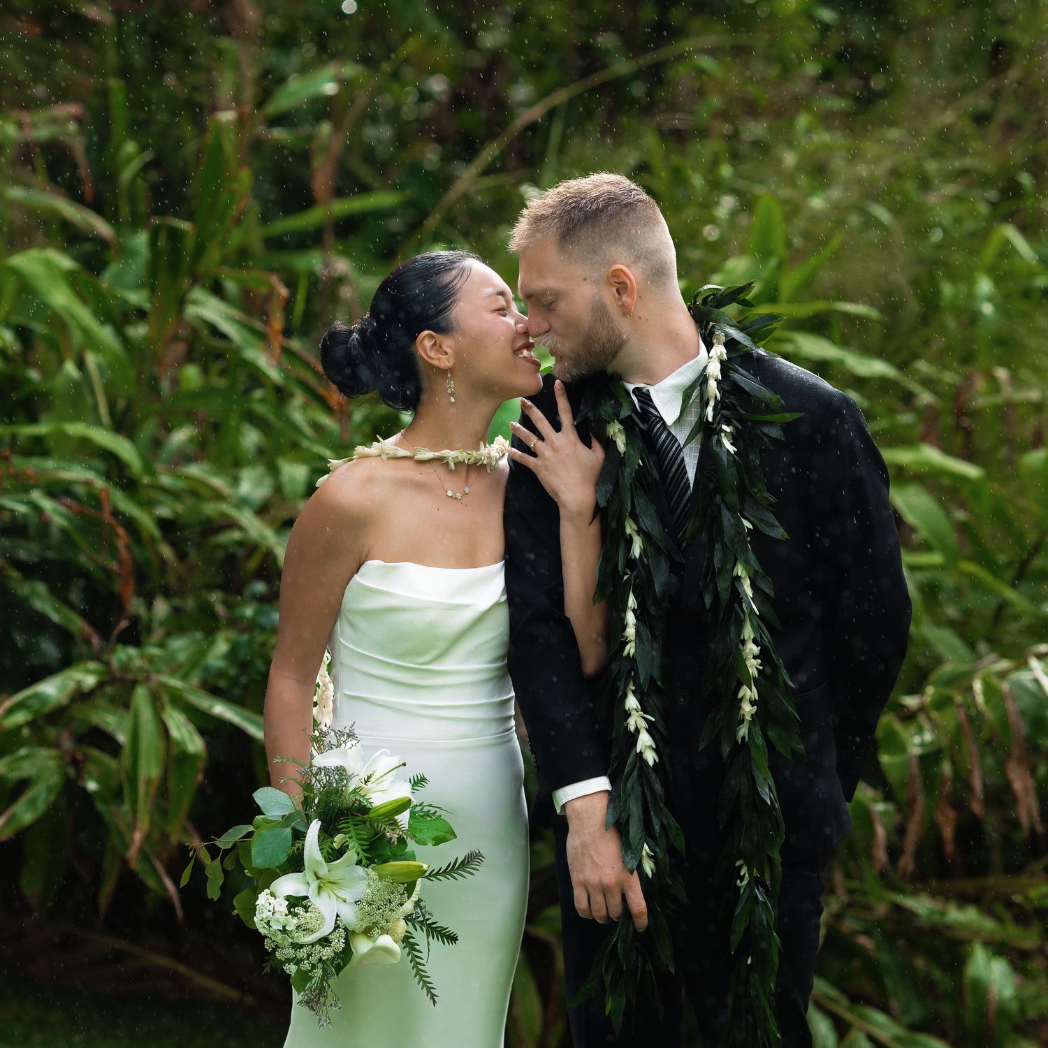 Couple sharing a romantic moment in lush tropical greenery, bride in white gown holding bouquet, groom wearing floral lei.