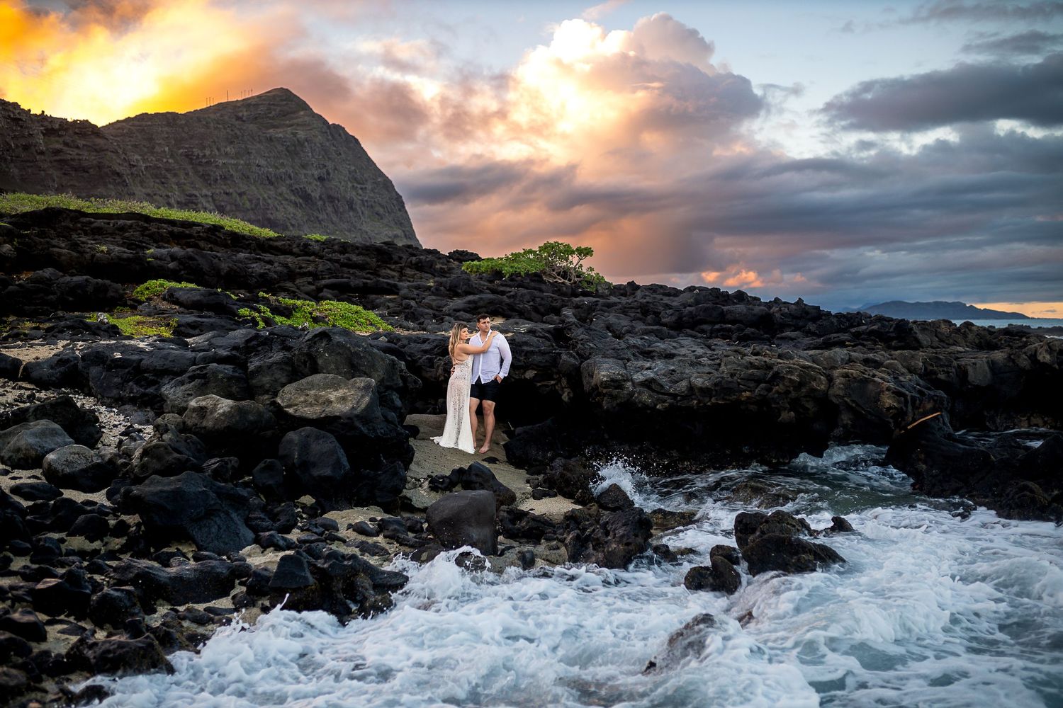Couple embracing on volcanic rocks at sunset in Hawaii, waves crashing around them under a dramatic colorful sky.