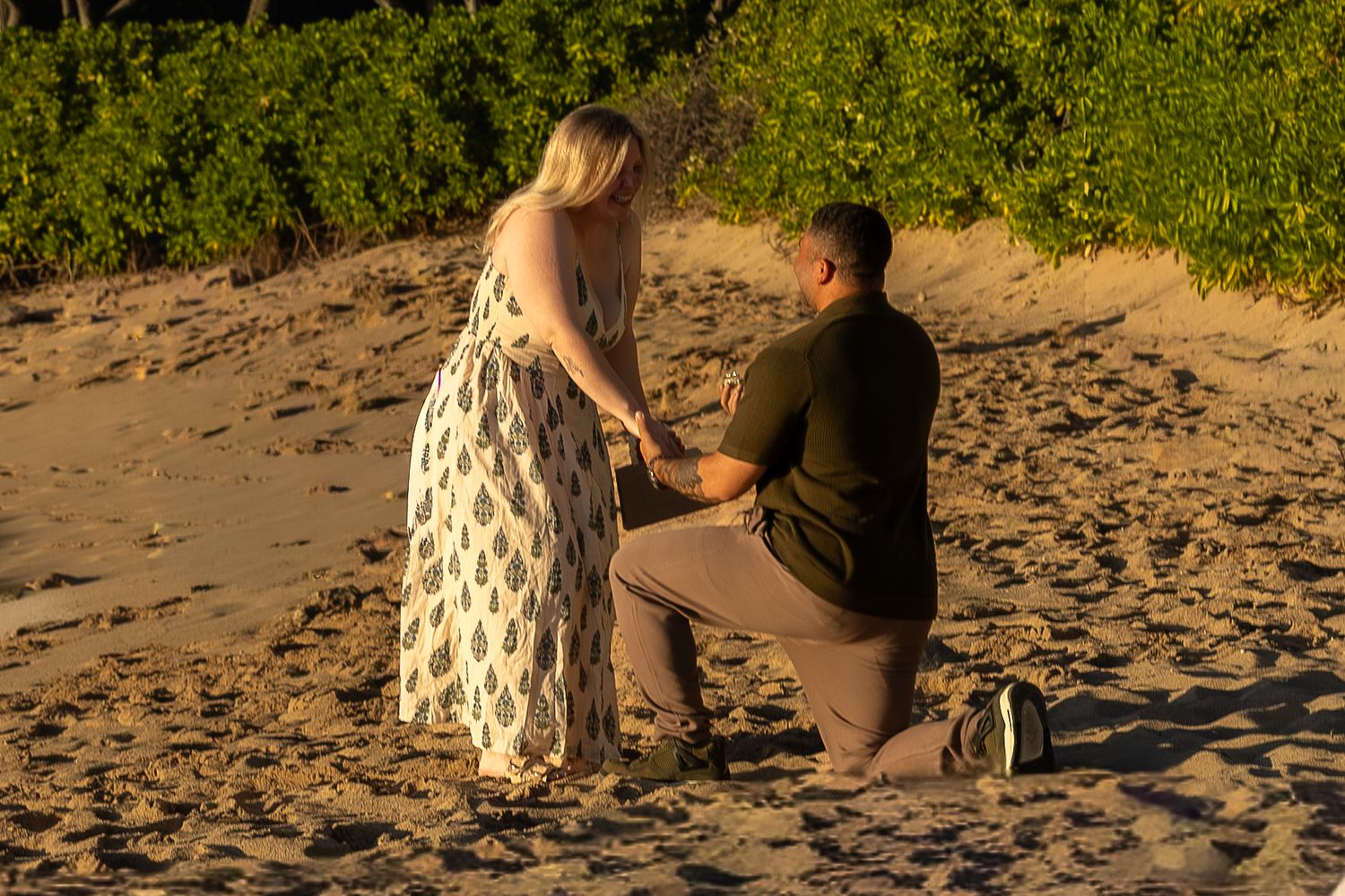 Man kneeling on sandy beach proposing to blonde woman at golden sunset near tropical greenery.