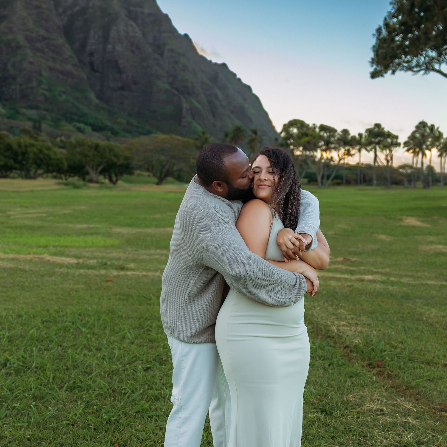 Joyful couple hugging and kissing on lush green field with scenic Hawaiian mountains.