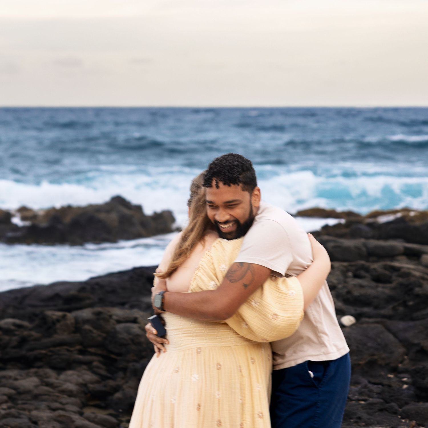 Newly engaged couple sharing joyful embrace on Hawaiian beach with dramatic rocky coastline and ocean waves.