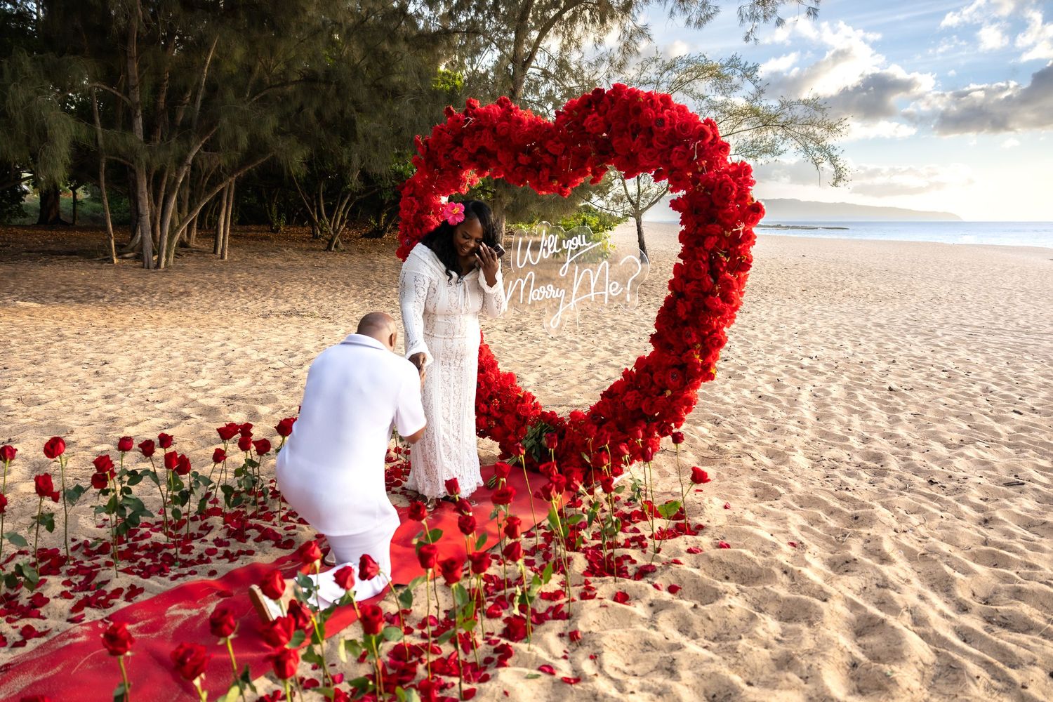 Man kneeling before woman during beach proposal surrounded by red roses and heart-shaped floral arch.