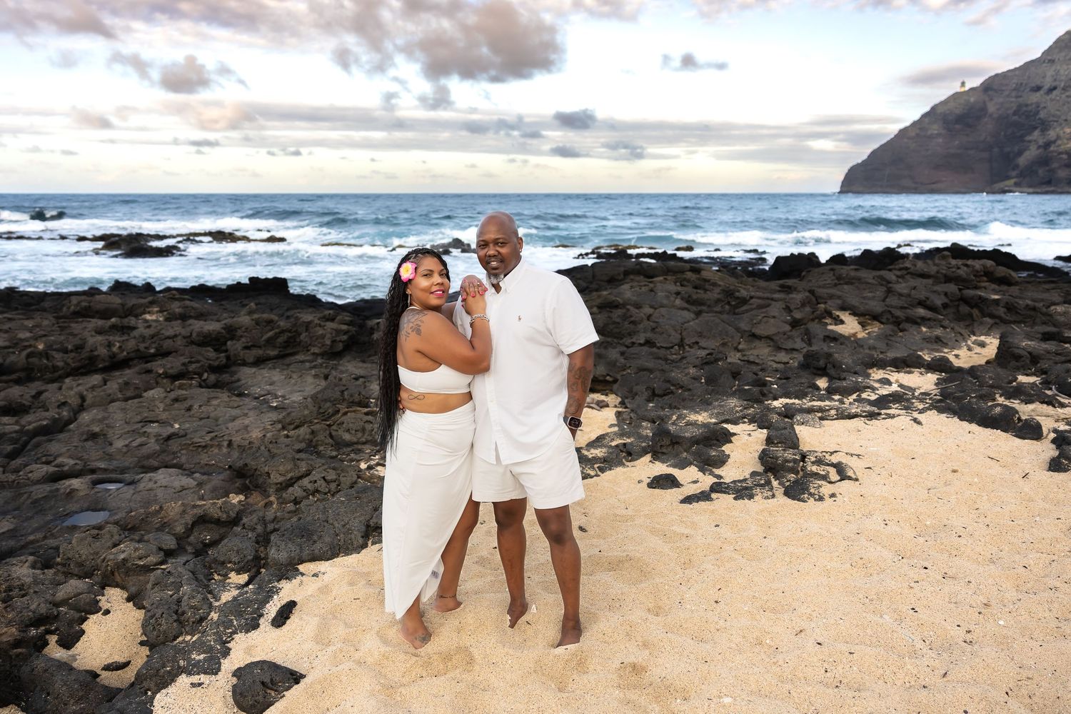 A couple in white outfits poses together on a sandy Hawaiian beach with rocky cliffs behind them.