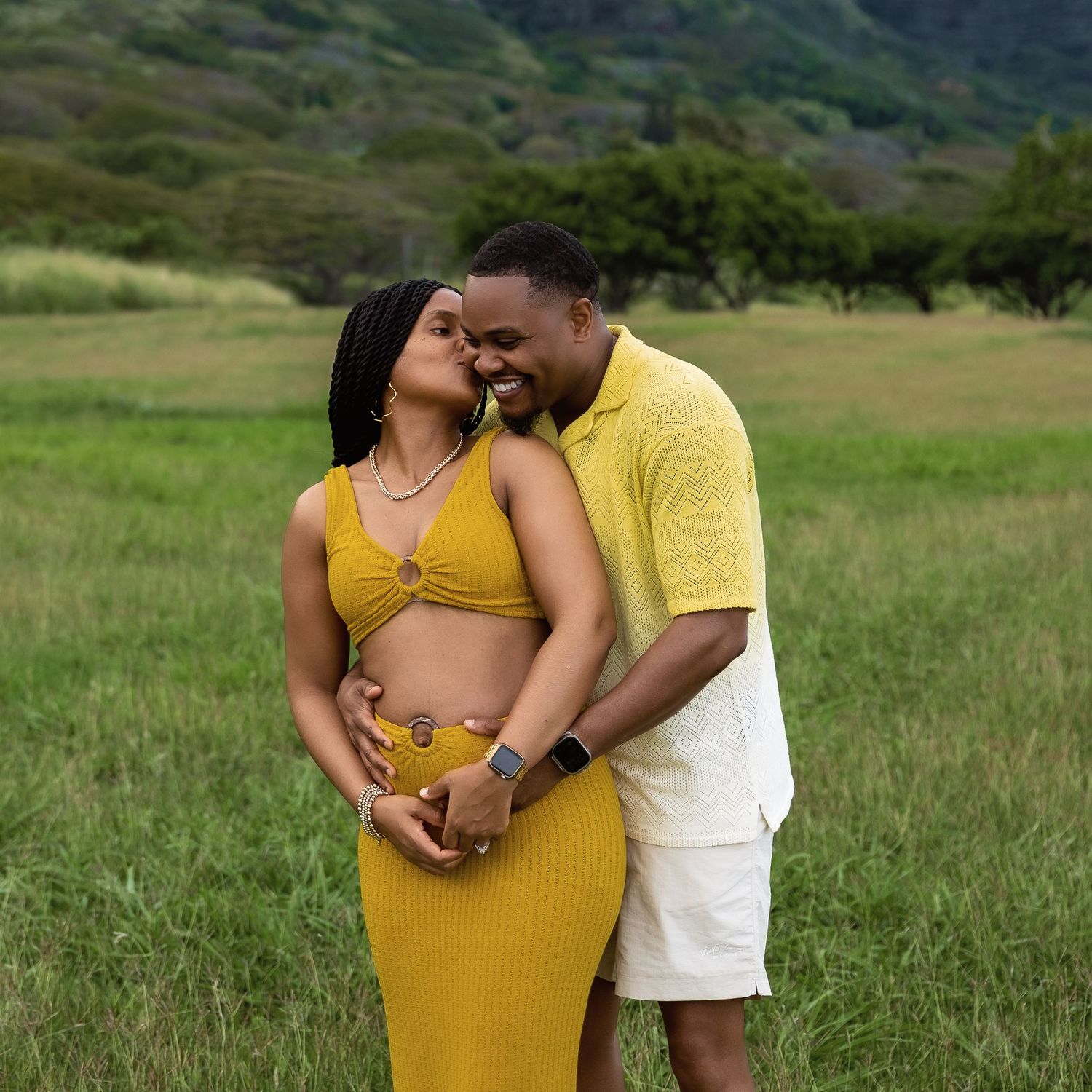 Expecting couple in matching yellow outfits embracing in a lush green Hawaiian valley field.