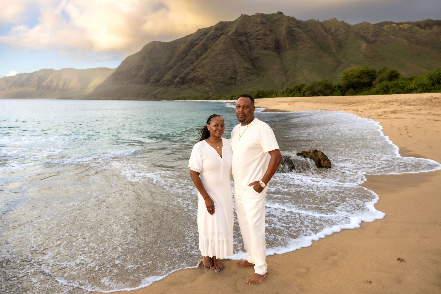 Couple in matching white outfits posing on a Hawaiian beach with mountain backdrop.