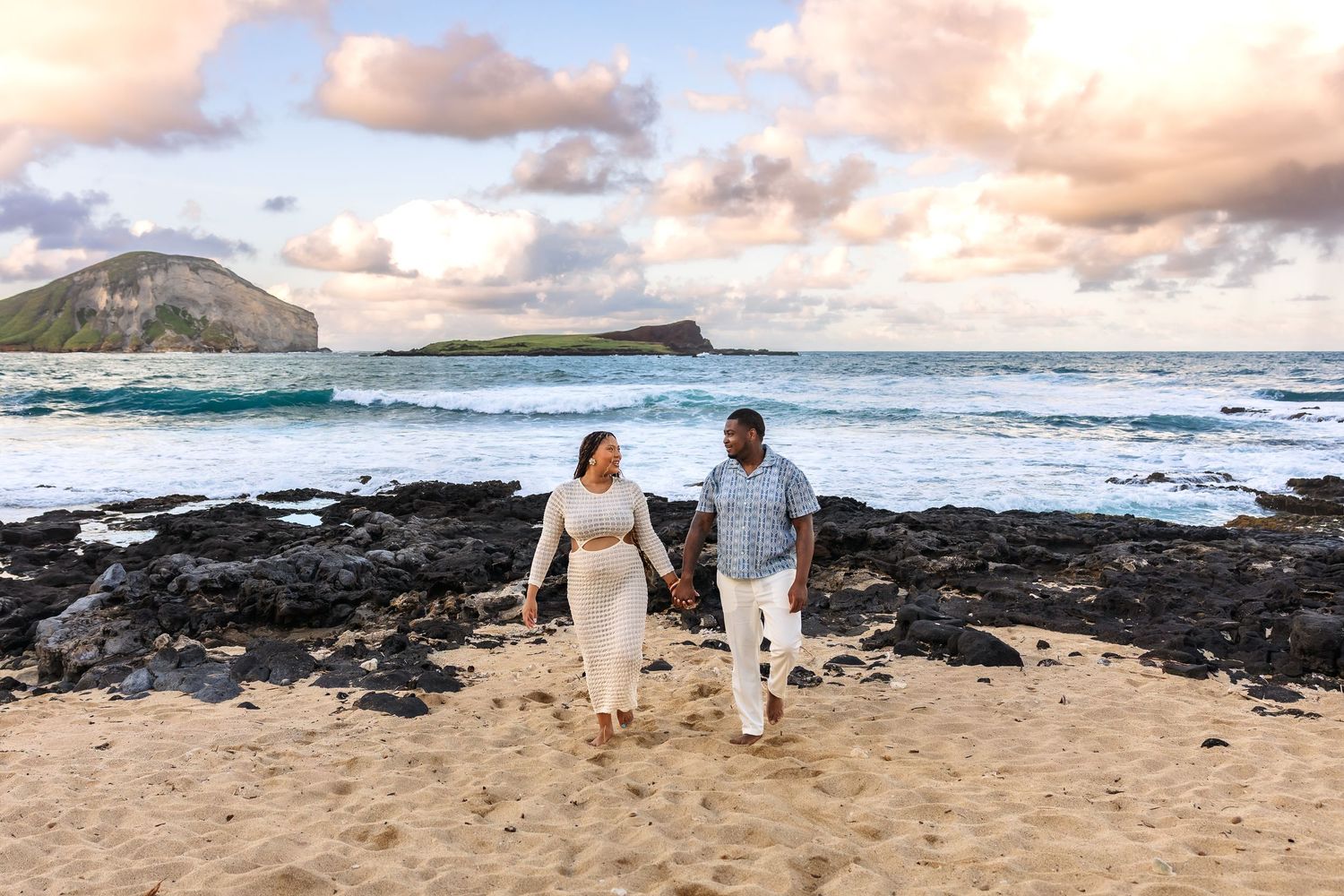 Expectant couple walks hand-in-hand along rocky Hawaiian beach at golden hour with ocean waves behind them.