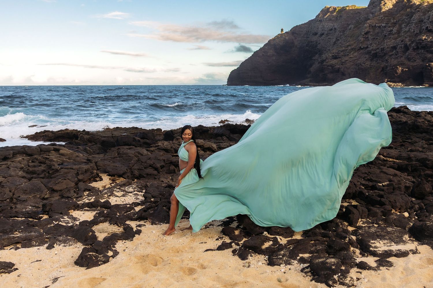 A woman in a flowing teal gown poses on rocky Hawaiian shoreline with ocean waves at sunset.