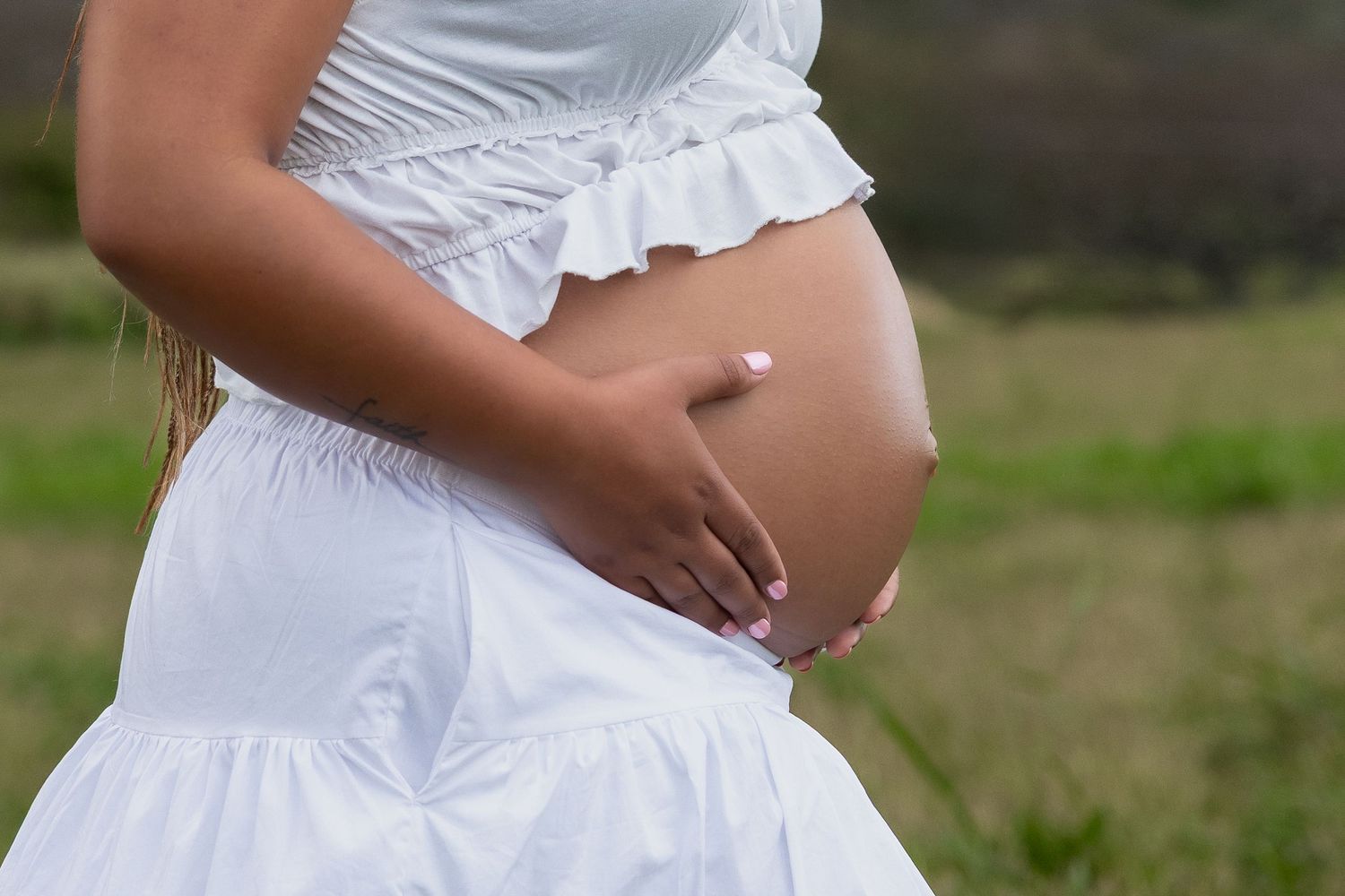 Pregnant woman in white dress cradling her baby bump outdoors in a green field.
