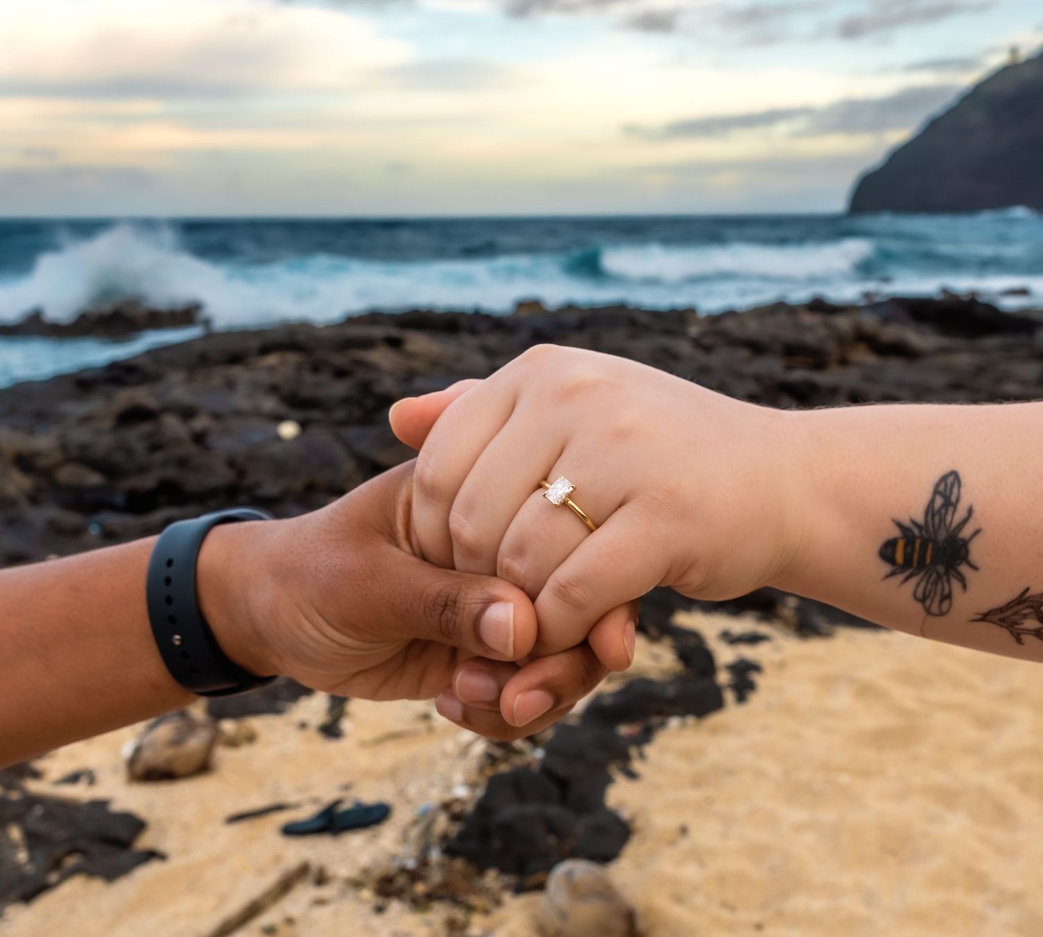 Close-up of interracial couple holding hands showing engagement ring, woman has bee tattoo on wrist.