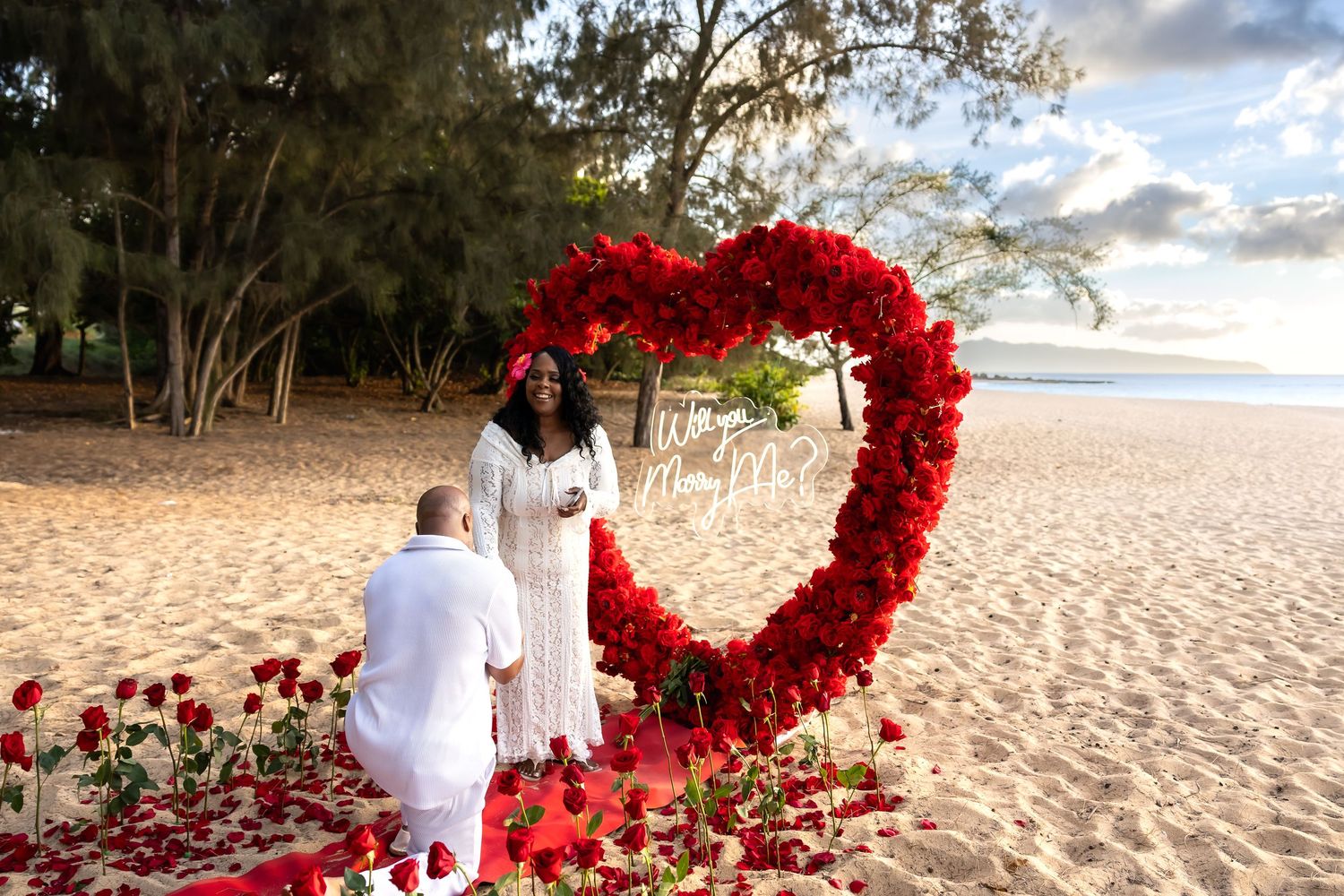 Romantic beach proposal with giant red rose heart arch and scattered rose petals at sunset.