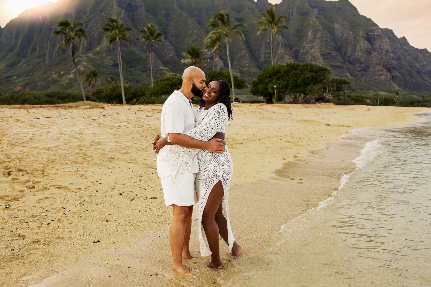 Couple embracing on a Hawaiian beach in white outfits with lush green mountains and palm trees in the background.