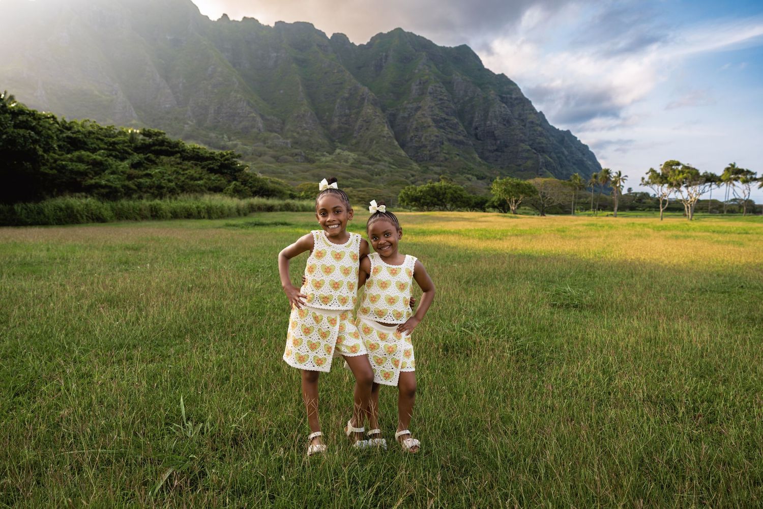 Two young girls in matching outfits posing in a green meadow with dramatic Ko'olau mountains behind.