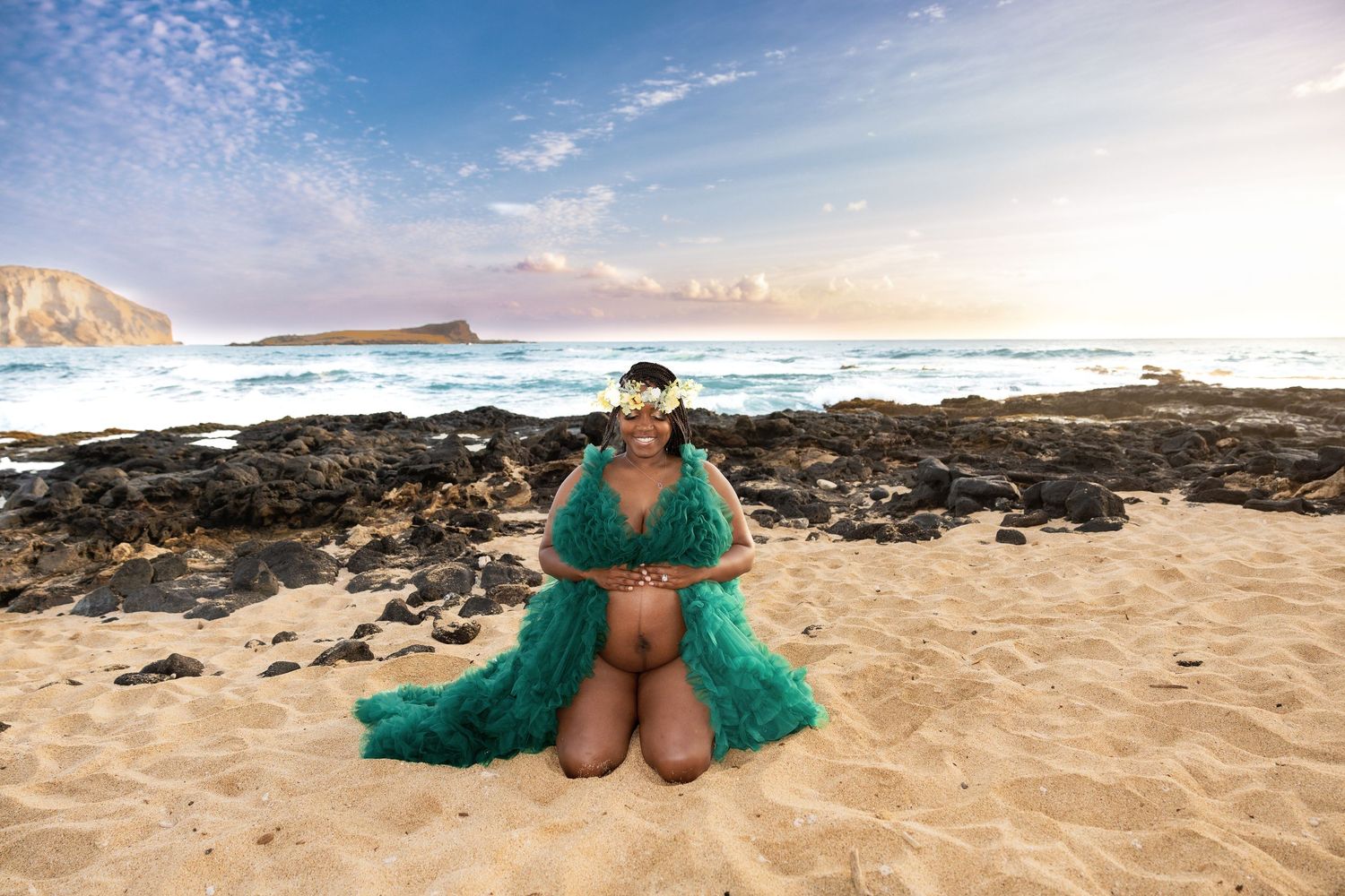 Pregnant woman in teal gown sitting on sandy beach with flower crown and ocean backdrop.