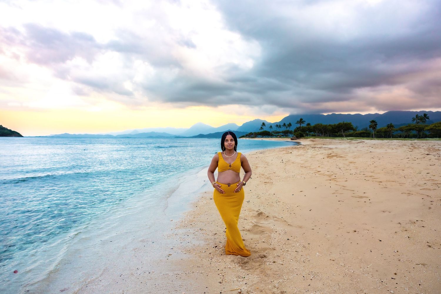 Pregnant woman in yellow dress poses alone on calm Hawaiian beach at sunset with mountains in the distance.