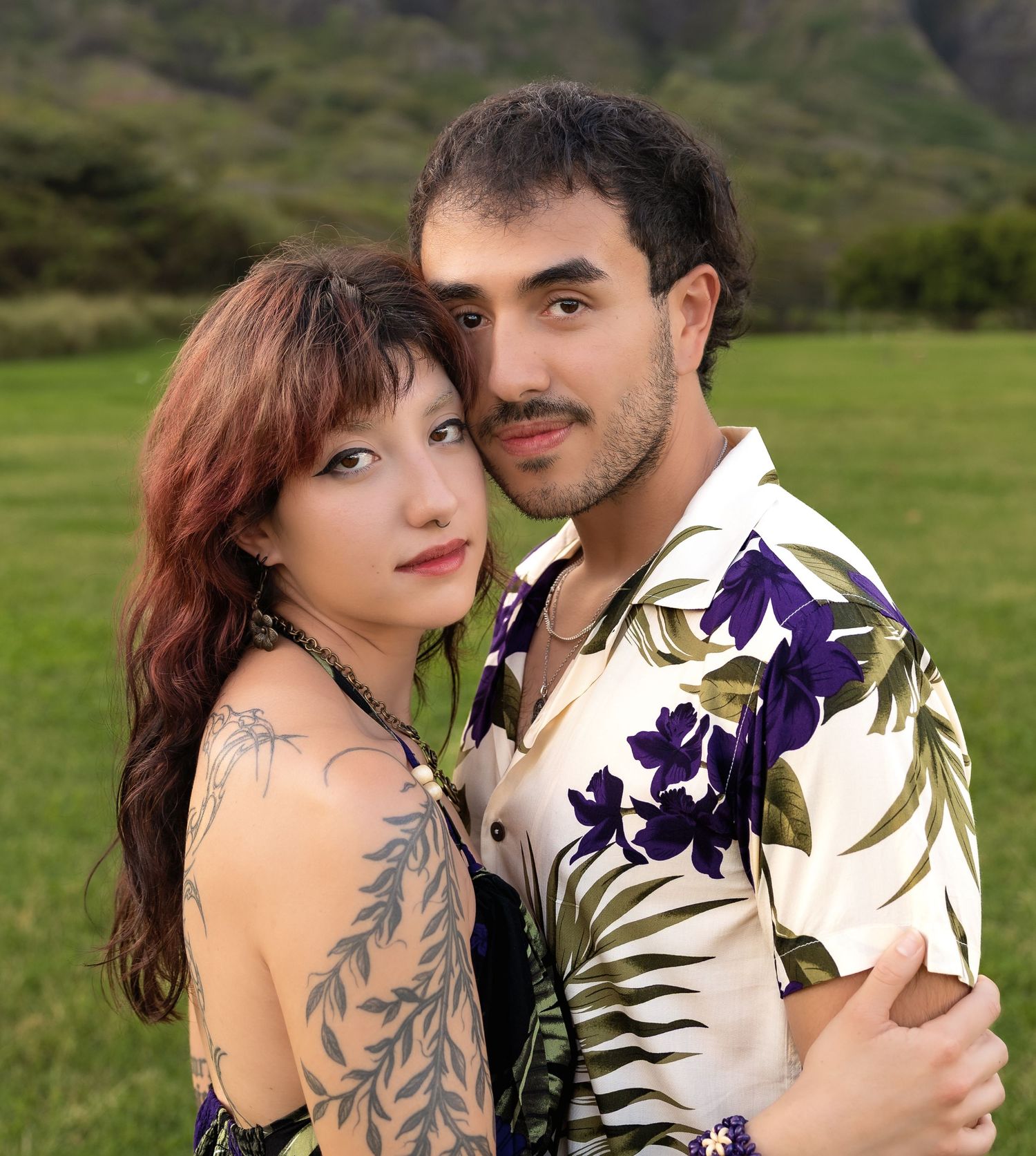 Young couple embracing in Hawaiian valley, woman with tattoos in floral dress, man in tropical shirt.