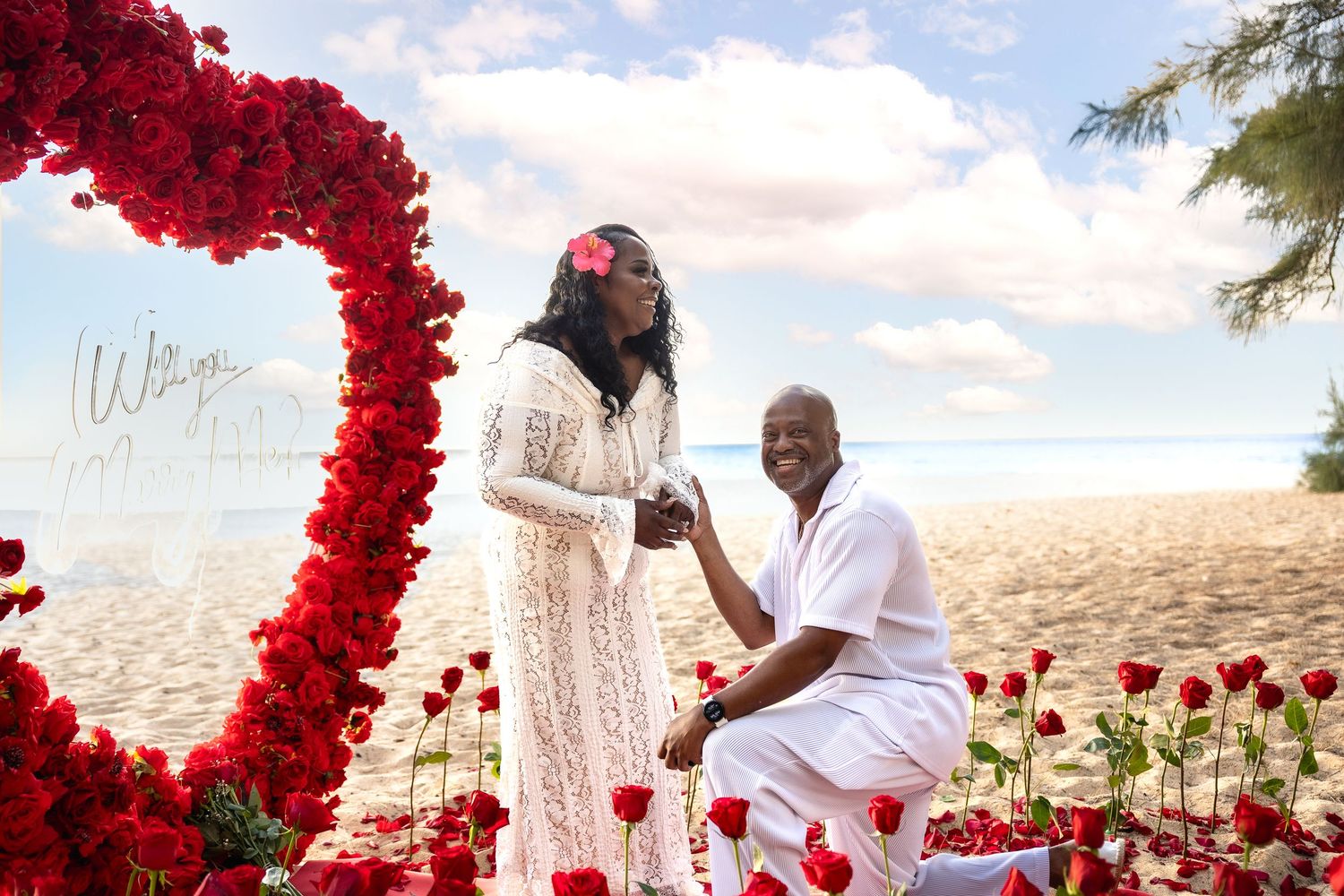 Man on one knee proposing to woman with engagement ring beside large red rose heart on beach.