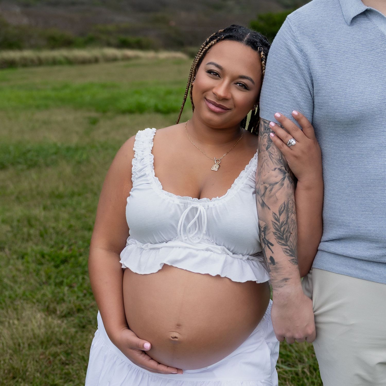 Pregnant woman in white crop top smiling outdoors during maternity photoshoot in green field.