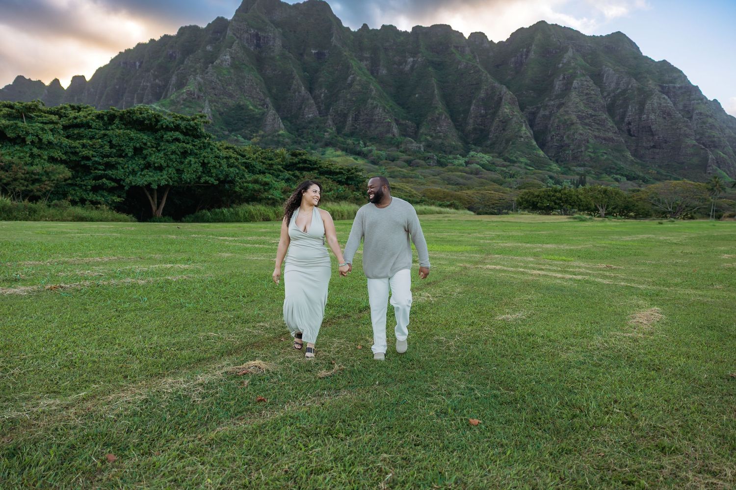 Happy couple holding hands walking through green Hawaiian meadow with majestic Ko'olau mountains behind them.