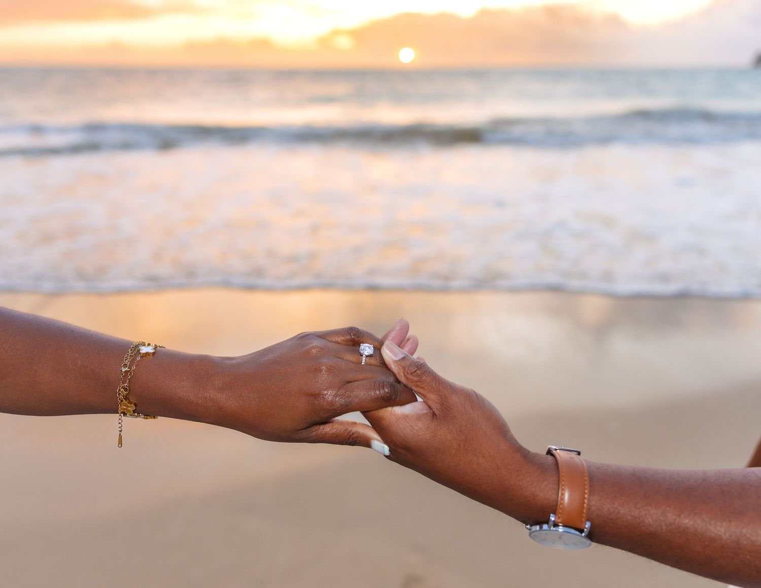 Two hands holding on a beach at sunset, one wearing a diamond ring and gold bracelet, the other a watch and bracelet.