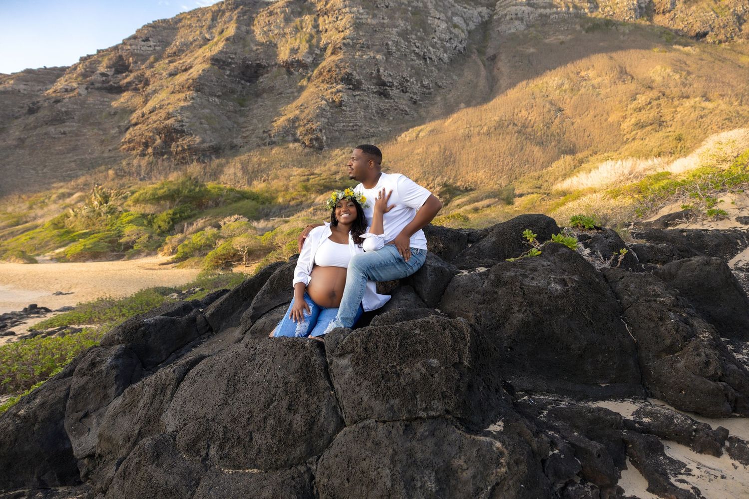 Couple sitting together on large rocks with scenic Hawaiian mountain and beach backdrop.