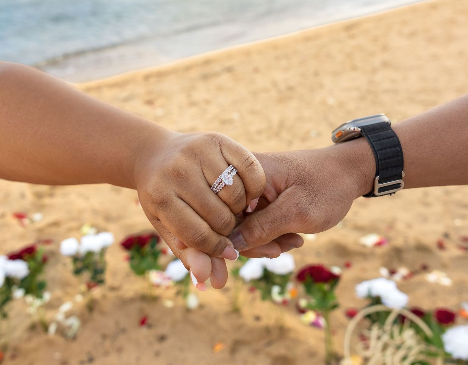 Close-up of couple holding hands on beach, woman wearing diamond engagement ring, man wearing black smartwatch.