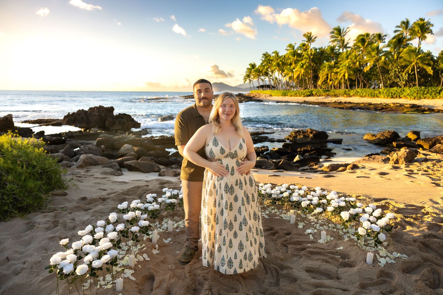Couple posing on a sandy beach surrounded by white flowers with palm trees at sunset.