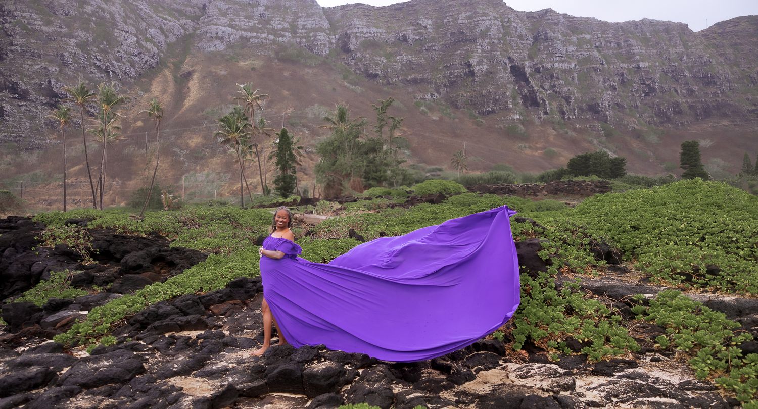 Woman holding a flowing purple fabric dress on a rocky hillside with green tea fields and mountains.