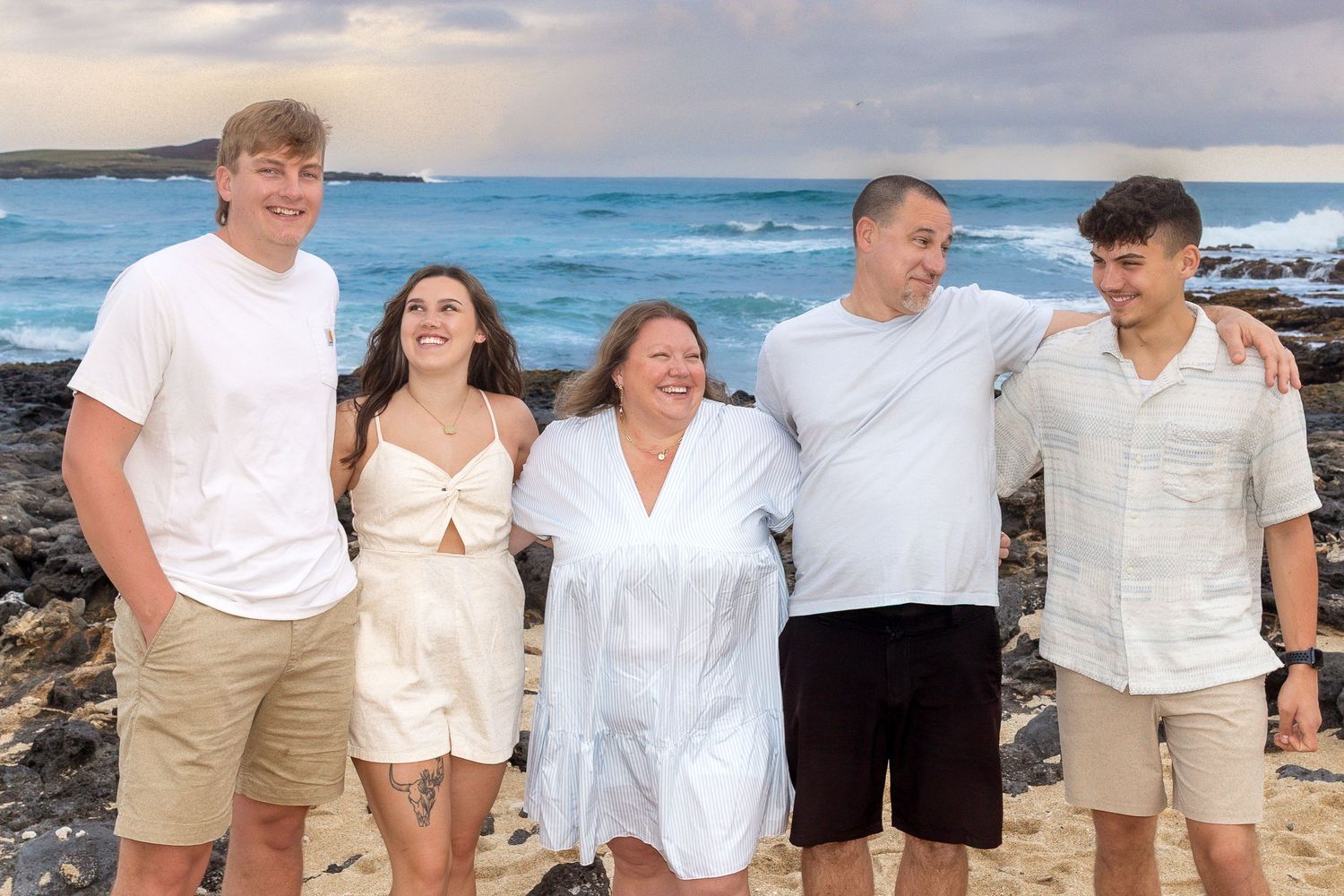 Family of five in white outfits smiles together on rocky Hawaiian beach with ocean waves behind them.