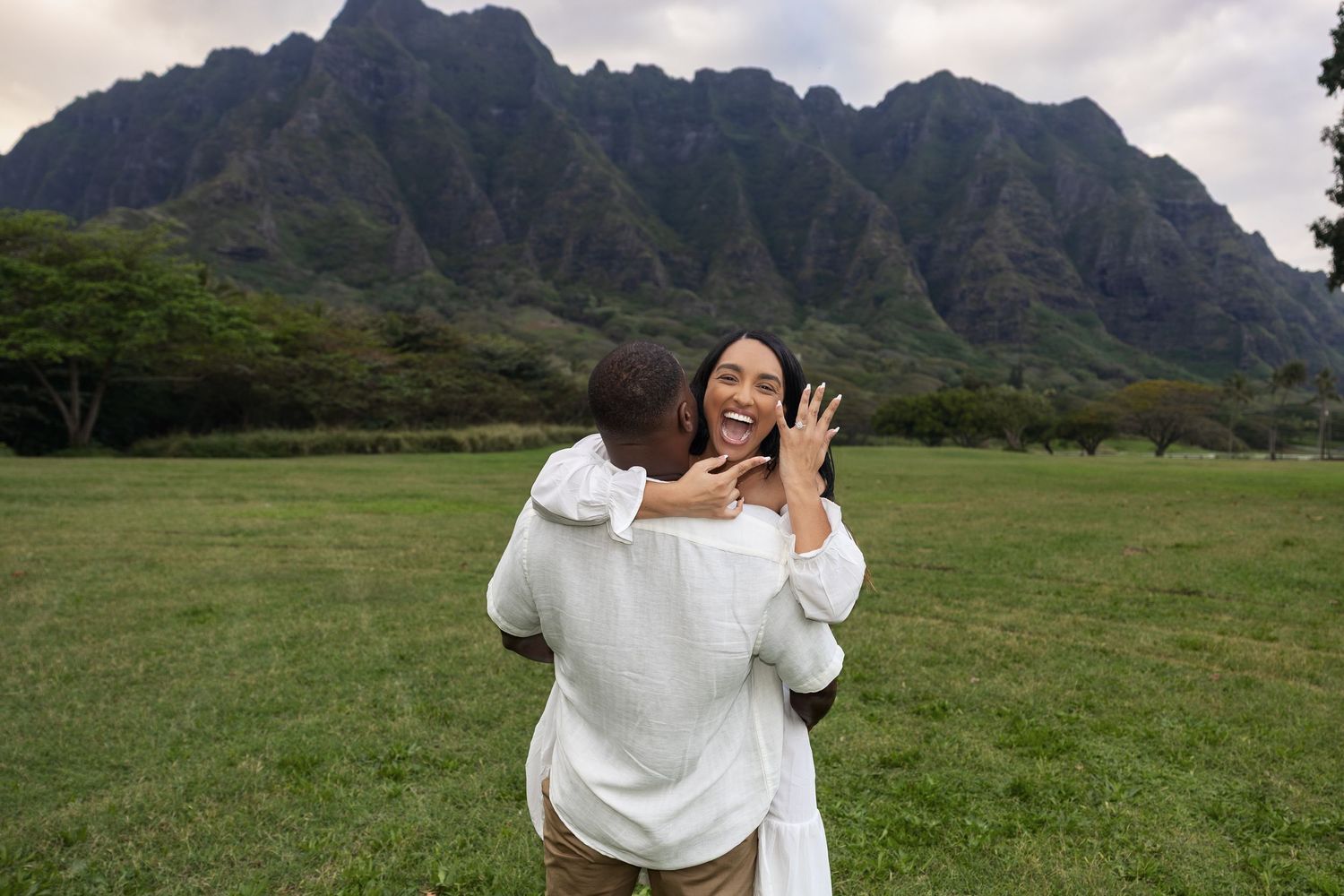 Man surprising woman with marriage proposal on green lawn with stunning mountain backdrop in Hawaii.