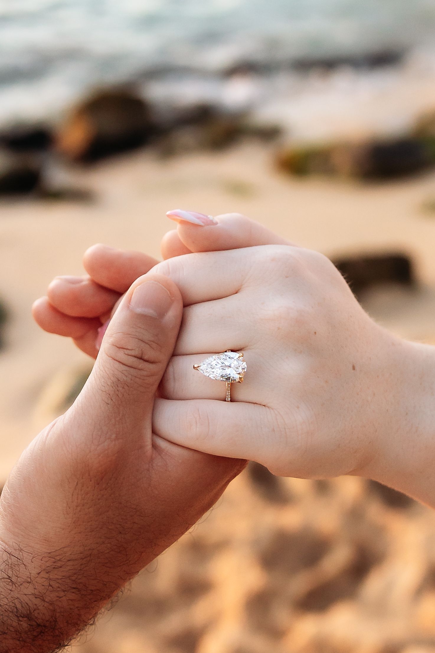 Close-up of hands showing a stunning pear-shaped diamond engagement ring on the beach.