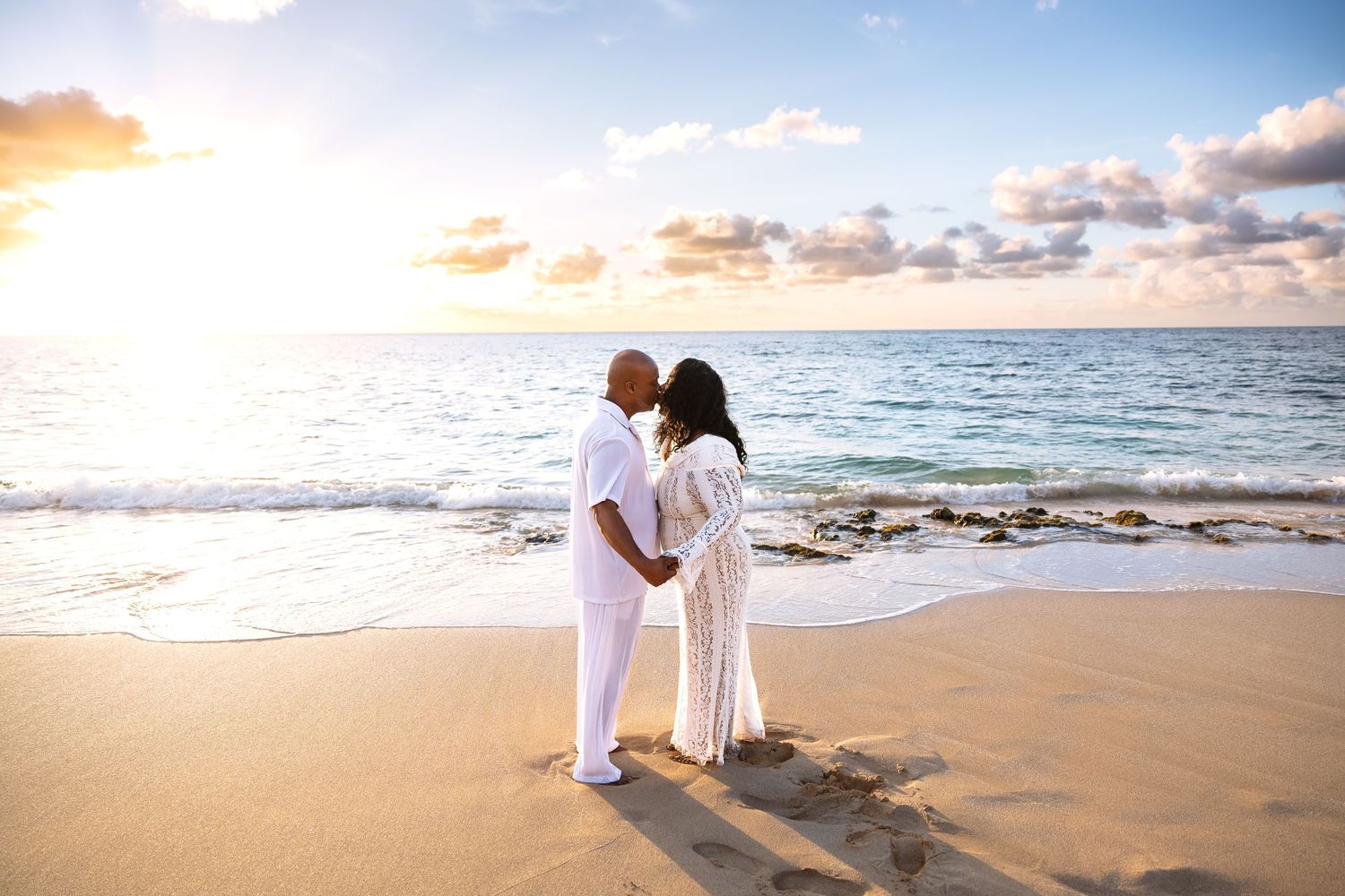 A couple in white holds hands facing each other on a Hawaiian beach during a golden sunset.