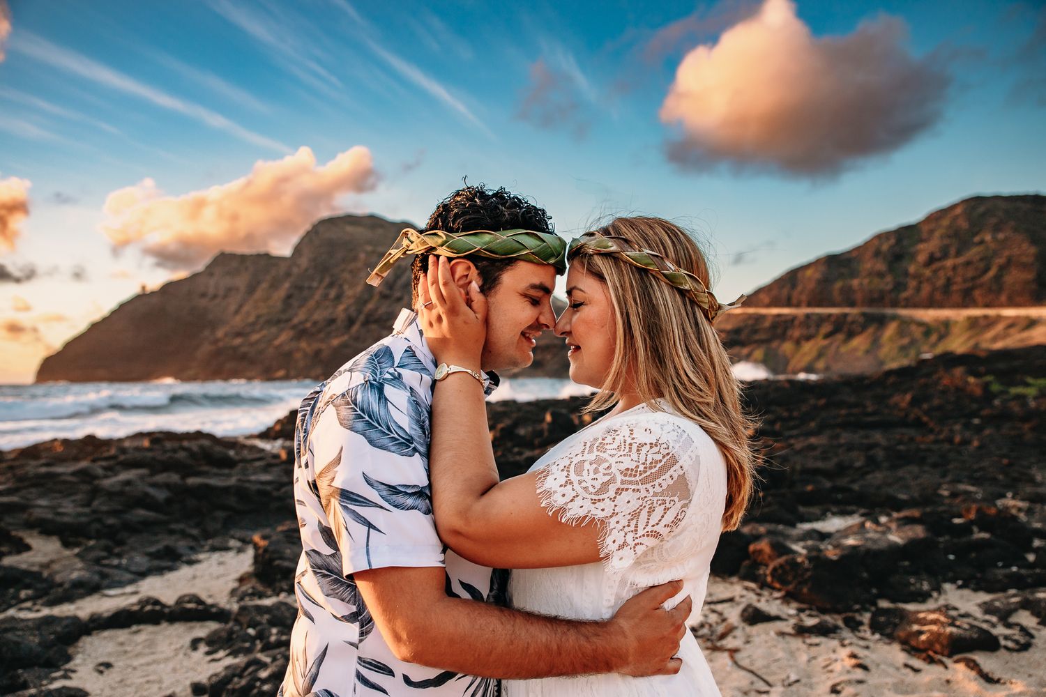Couple sharing a romantic moment on a rocky Hawaiian beach at sunset with mountains in the background.