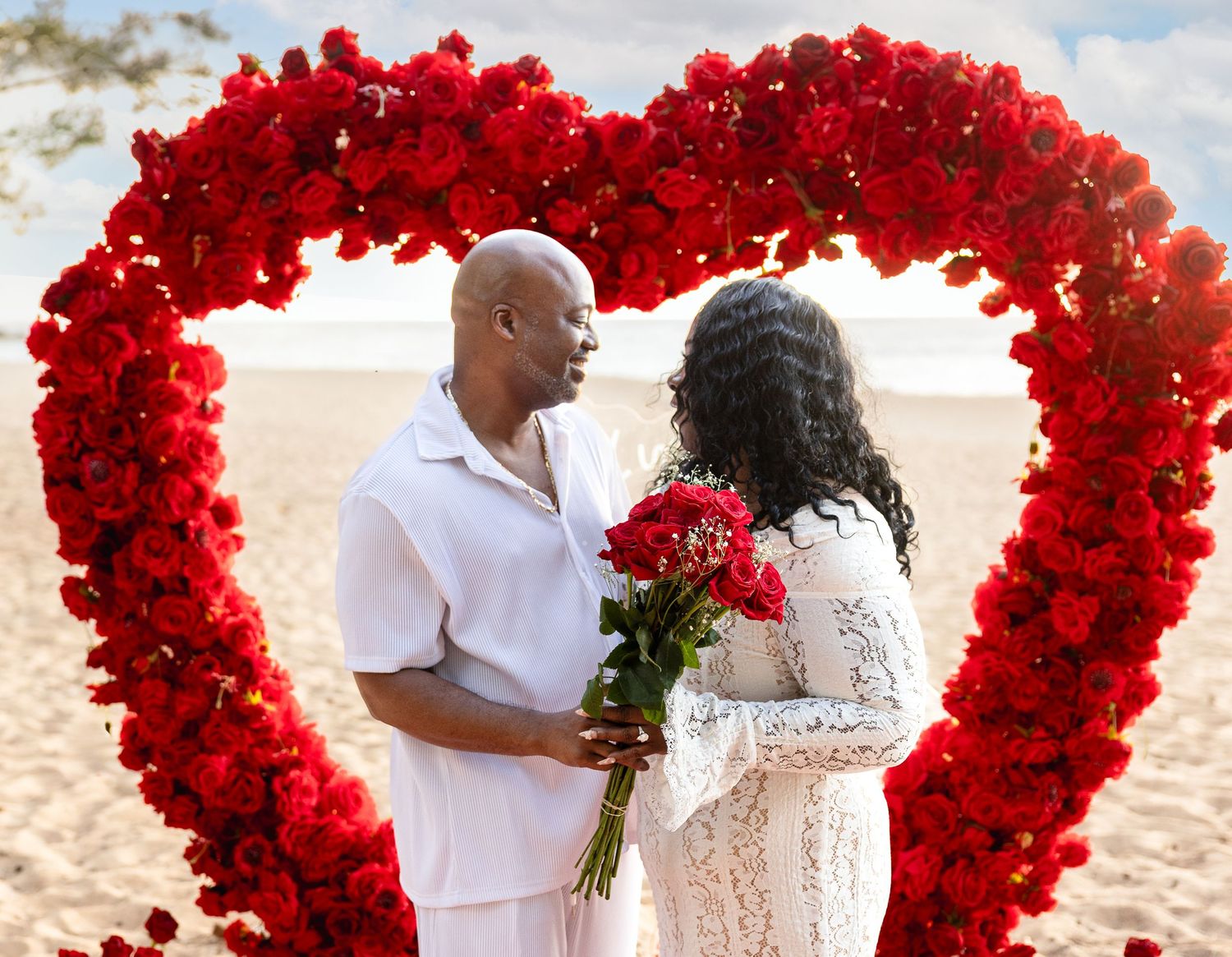 Happy couple standing inside stunning red rose heart arch on tropical beach after proposal.