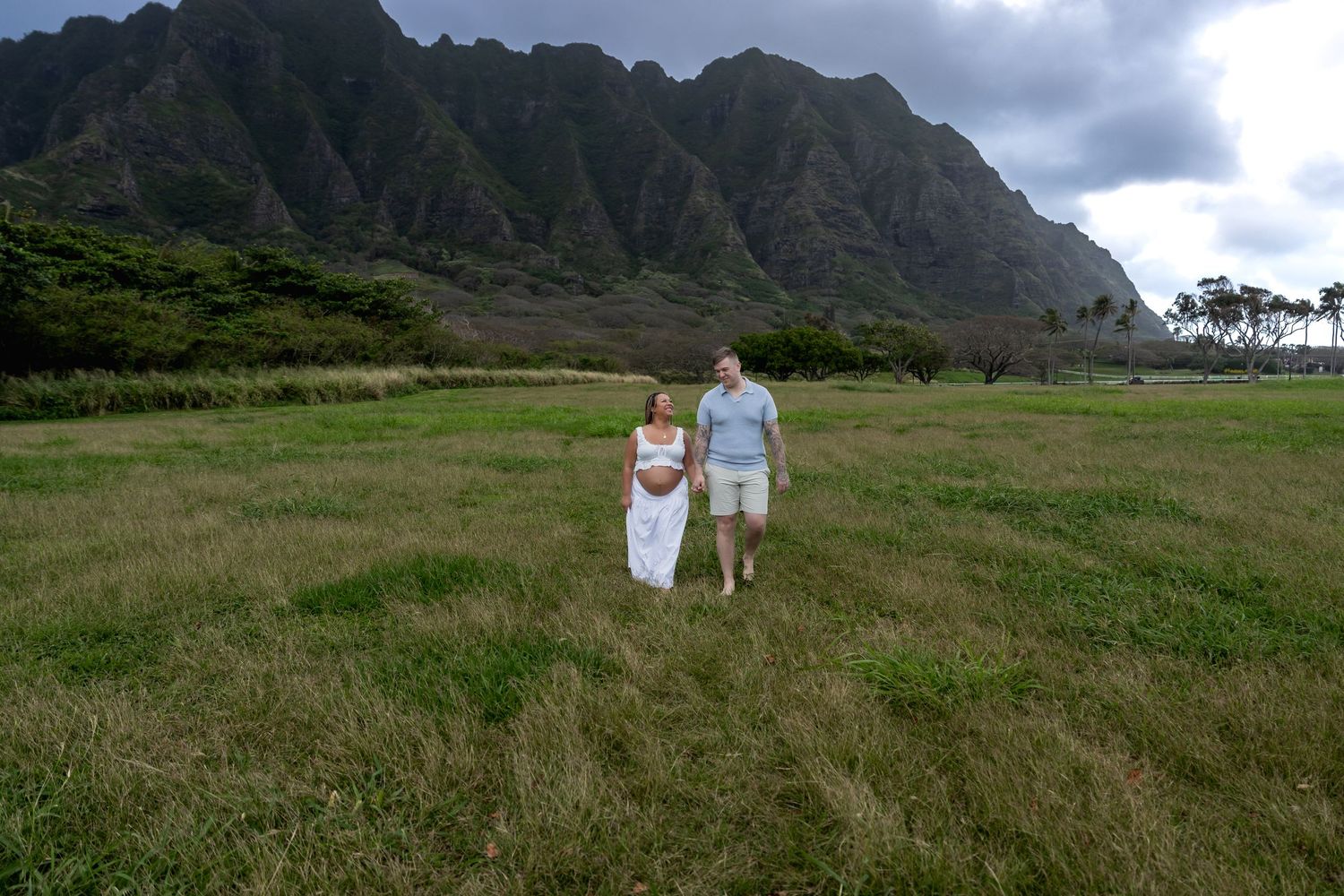 Couple walking hand in hand through green field with dramatic mountain backdrop in Hawaii.