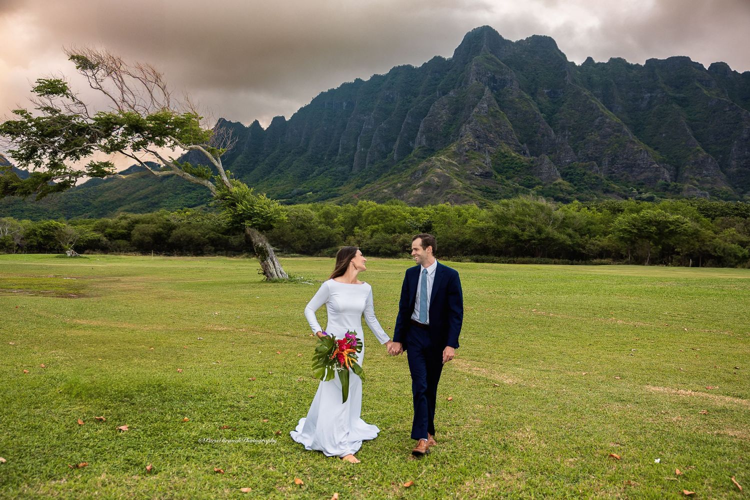 Newlyweds walking hand in hand across a green meadow with the stunning Ko'olau mountains in Hawaii.
