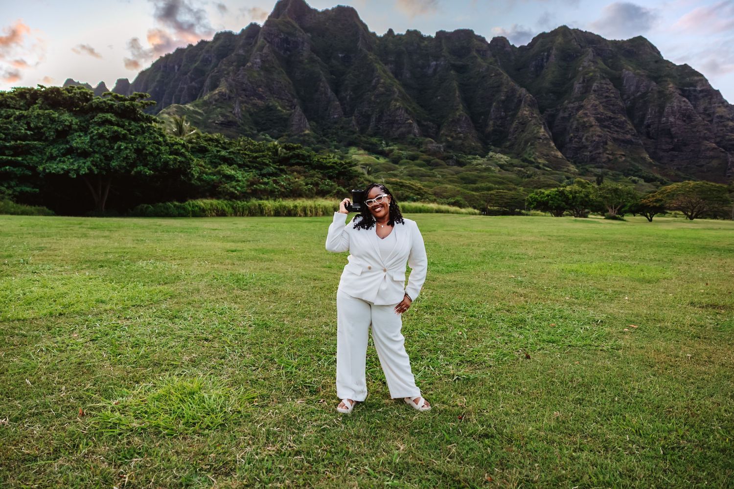 Woman in white outfit laughs joyfully in green field with dramatic Ko'olau mountain backdrop in Hawaii.
