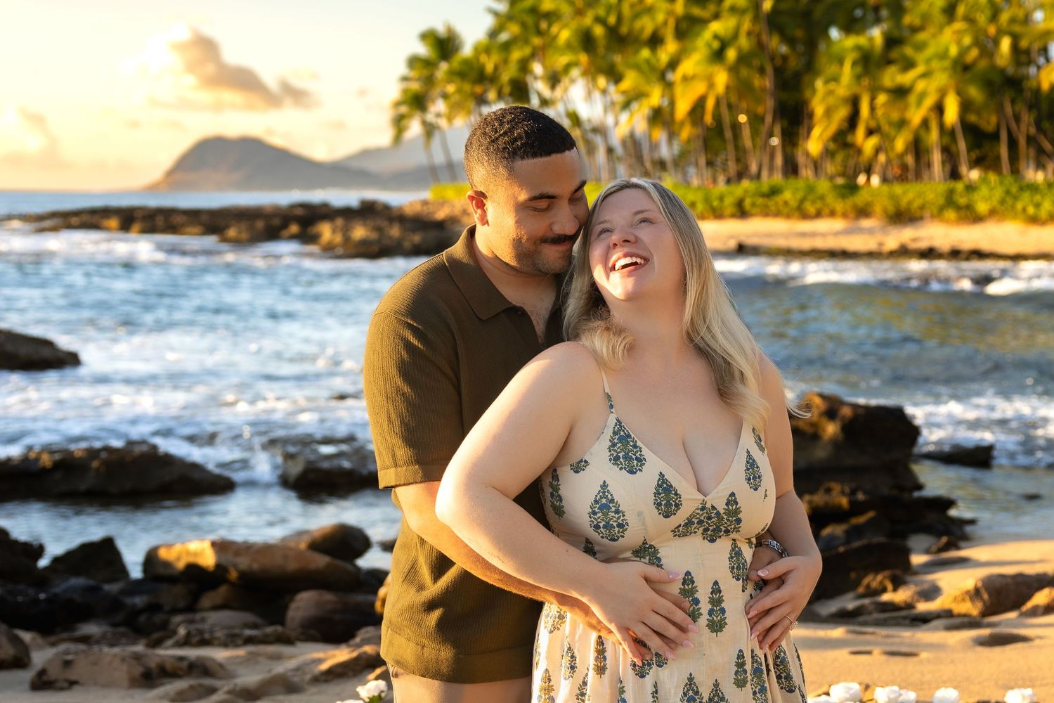 Happy couple embracing on a Hawaiian beach at sunset with mountain views and palm trees.
