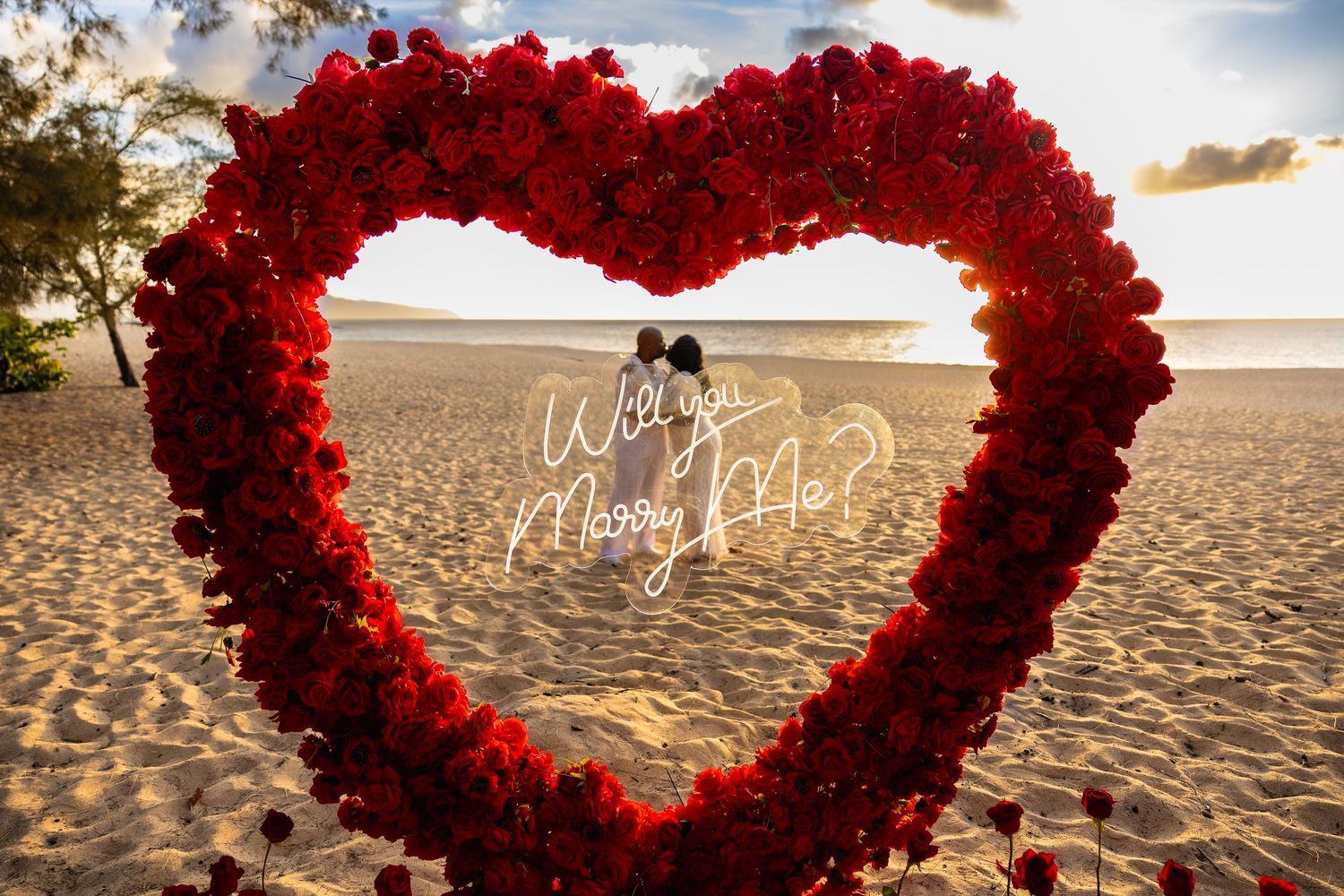 Will you marry me neon sign inside giant red rose heart arch on romantic sunset beach.