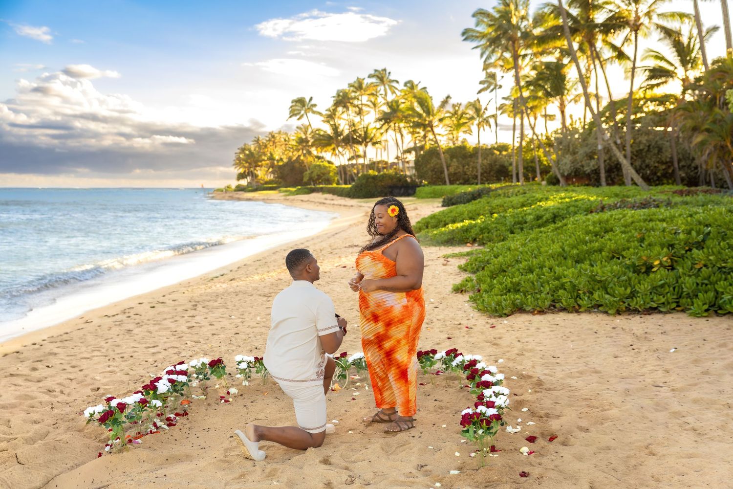 Man proposing on knee to woman in orange dress within heart-shaped floral arrangement on beach.