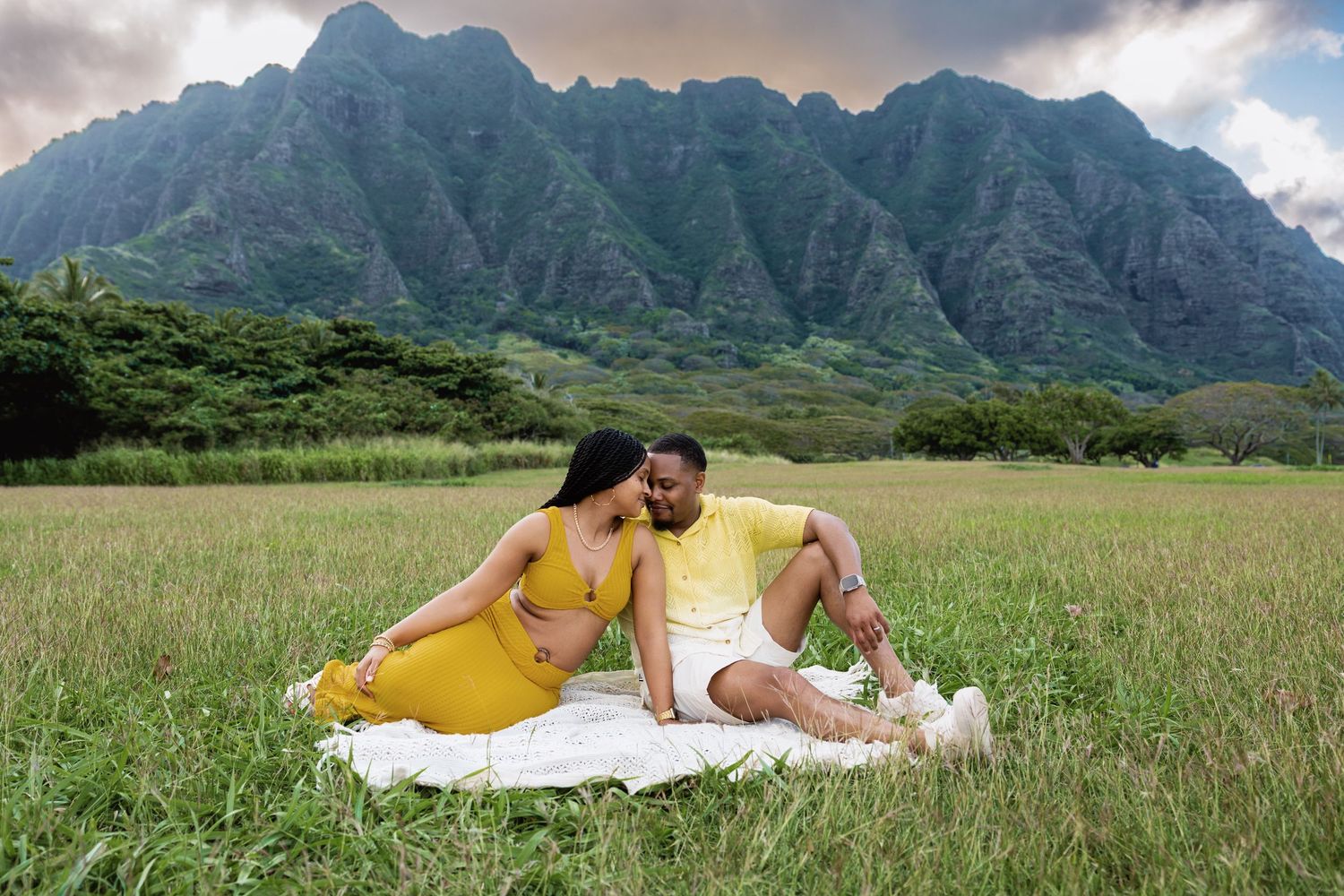 Couple sits together on white blanket in lush Hawaiian meadow with stunning Ko\u02bbolau mountain range backdrop.