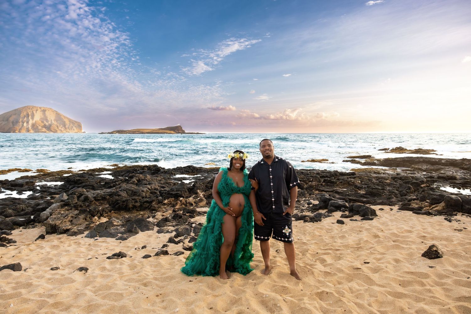 Couple posing together on rocky Hawaiian beach at golden sunset with ocean island backdrop.
