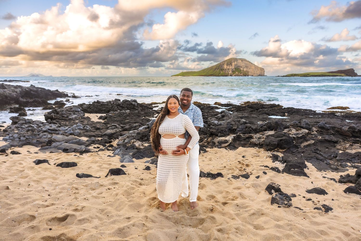 Couple embraces on Hawaiian beach at sunset, man holds pregnant partners belly with rocky coast and island backdrop.
