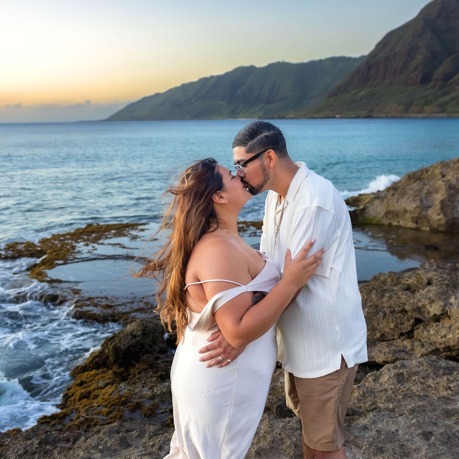 Romantic couple embracing and kissing on rocky Hawaiian coastline at sunset.