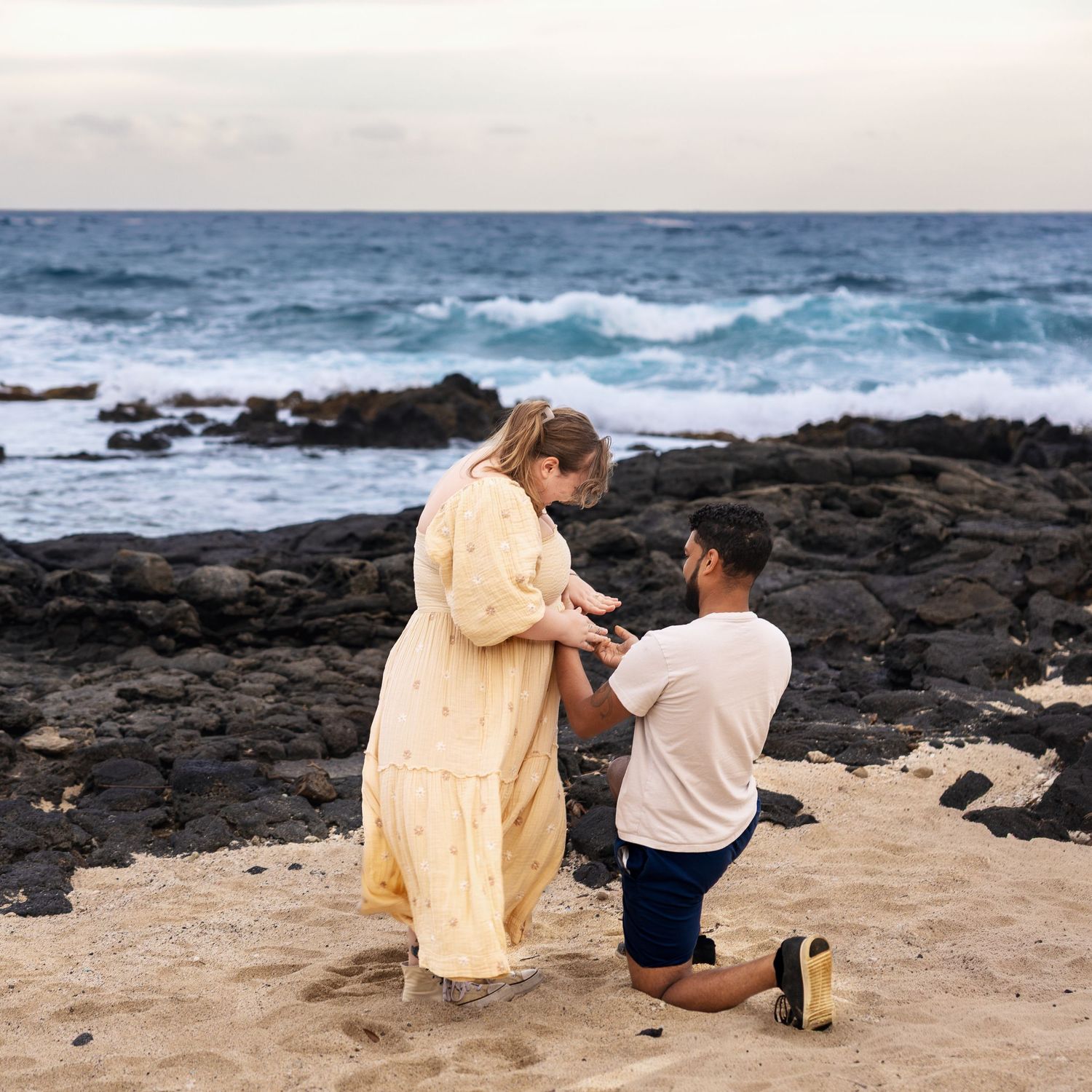 Man proposing on one knee to woman in yellow dress on sandy Hawaiian beach with rocky coastline and waves.