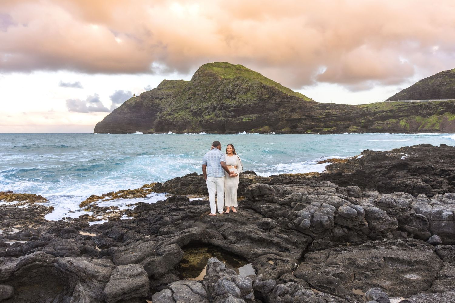 Couple stands together on rocky Hawaiian lava coastline gazing at turquoise ocean and lush green mountain at sunset.