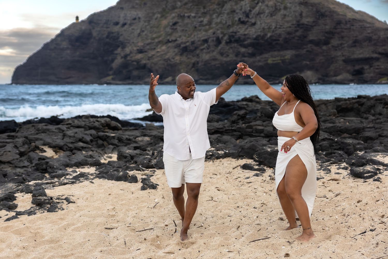 A couple dances playfully on a Hawaiian beach with rocky shoreline and ocean waves behind them.