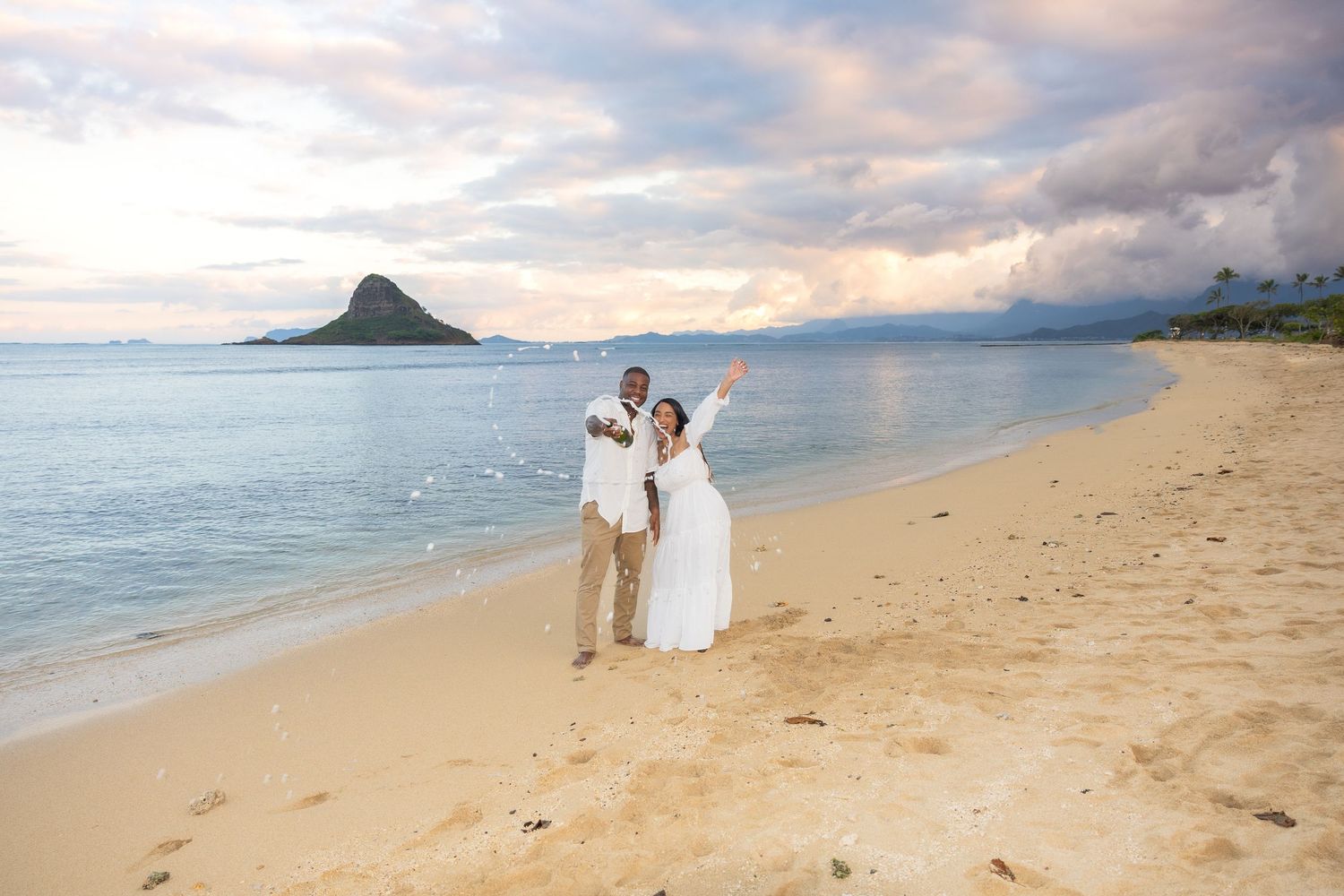 Happy couple in wedding attire celebrates on sandy Hawaiian beach with Mokoli'i Island in background.