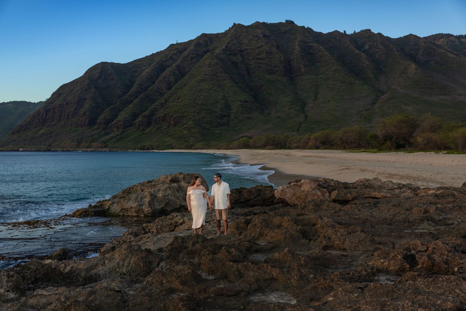 Couple standing on rocky shore with stunning mountain backdrop and turquoise ocean at Hawaiian beach sunset.