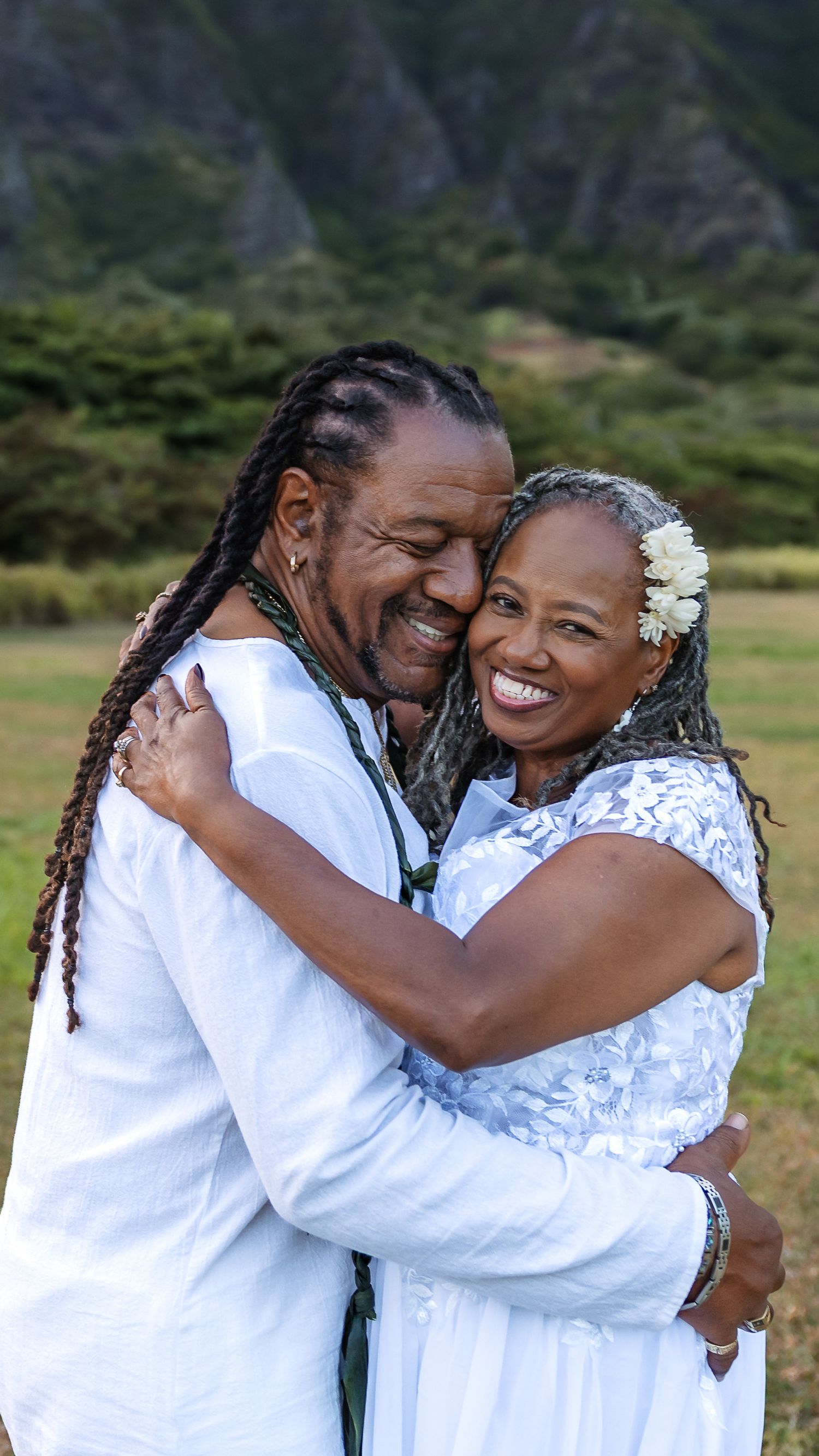 Joyful mature couple in white embracing and laughing with lush Hawaiian mountain greenery behind them.